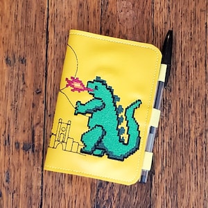 May include: A yellow leather notebook with a green pixelated monster design. The monster is breathing fire and there is a black pen next to the notebook.