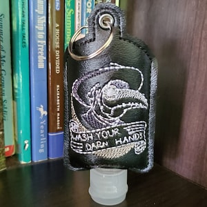 May include: Black leather hand sanitizer holder with a silver key ring. The holder features an embroidered design of a skull wearing a hat and the words "WASH YOUR DARN HANDS". The holder is attached to a clear plastic bottle.