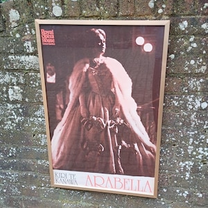 May include: A vintage poster for the opera Arabella, featuring a woman in a white gown and fur stole, with the text "Royal Opera House Covent Garden" and "Kiri Te Kanawa" at the top and bottom of the poster.