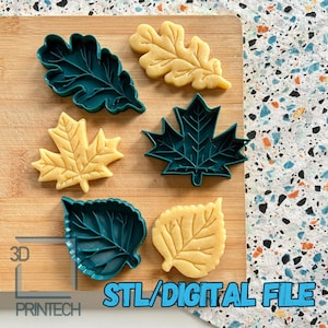 May include: Set of six 3D printed leaf cookie cutters. The cutters are green and feature different leaf shapes, including oak, maple, and birch. The cutters are shown on a wooden surface with a light brown background.
