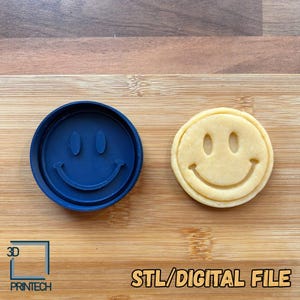 May include: A dark blue circular cookie cutter featuring a smiley face design with two oval eyes and a curved mouth, positioned next to a matching light beige circular cookie with the same embossed smiley face design. Text reads 3D PRINTECH STL/DIGITAL FILE.