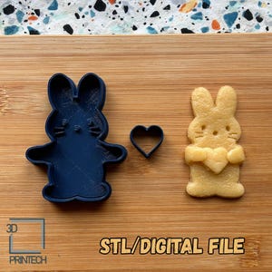 May include: A dark blue bunny-shaped cookie cutter, a small heart-shaped cutter, and a baked bunny cookie holding a heart. The text "STL/DIGITAL FILE" is printed in orange. The cookie cutter is on a wooden surface.