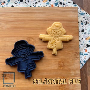 Op de afbeelding: Een blauwe plastic koekjesvorm in de vorm van een vogelverschrikker naast een gebakken vogelverschrikkerkoekje. De tekst "STL/DIGITAL FILE" staat onderaan de afbeelding, op een houten plank.