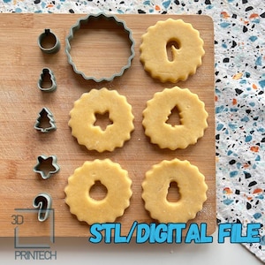 Op de afbeelding: Een set van 3D-geprinte kerstkoekjesvormen op een houten snijplank met verschillende koekjes die met de vormen zijn uitgestoken. De vormen omvatten een krans, een snoepstok, een sneeuwpop, een kerstboom en een ster. De tekst "STL/DIGITAL FILE" is zichtbaar in de rechterbenedenhoek van de afbeelding.