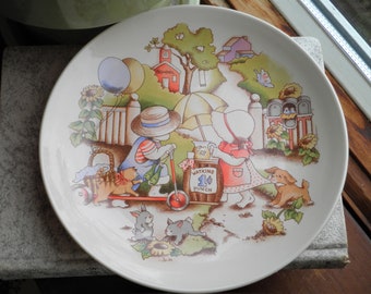 May include: A white ceramic plate with a colorful illustration of children playing. The scene includes a red schoolhouse, a lemonade stand, and a blue and white striped umbrella. The plate has a scalloped edge and is decorated with a floral pattern.