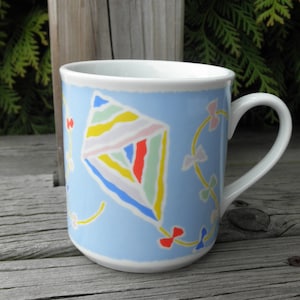 May include: A light blue ceramic mug with a colorful kite design. The kite is made up of yellow, red, green, pink, and blue stripes. The mug has a white handle.