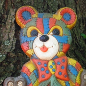 May include: A colorful patchwork teddy bear with a red bow tie. The bear is made of different colored fabric pieces sewn together. The bear has a white face with black eyes and a pink nose.
