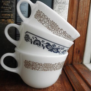 May include: Three white ceramic mugs with gold and blue floral designs stacked on top of each other. The mugs are sitting on a wooden surface.