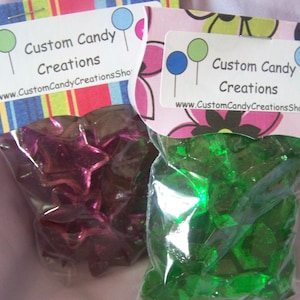 May include: Two bags of candy. One bag contains purple star-shaped candies. The other bag contains green, clear, irregularly shaped candies. Both bags have labels that say "Custom Candy Creations" and a website address.