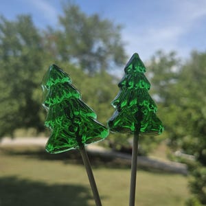 May include: Two green, tree-shaped lollipops on wooden sticks. The transparent candy has a detailed texture, resembling a miniature Christmas tree. The background features a blurred view of green trees and a blue sky.
