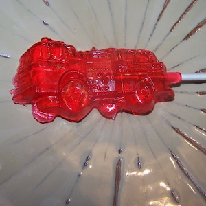 May include: A red candy lollipop shaped like a fire truck. The lollipop is on a white stick and is sitting on a white plate with a brown starburst design.