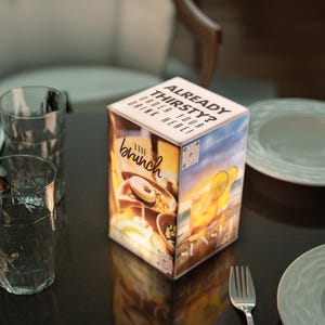 May include: A cube-shaped menu display with the text "ALREADY THIRSTY? ORDER YOUR DRINK HERE!" on top. The sides feature images of food and drinks, including a cocktail and the words "The Brunch" and "Sunset".