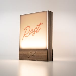 May include: A wooden lamp with a rectangular, illuminated front panel. The word "Rast" is written in coral script on the front. The base of the lamp is also wood, with the word "Rast" etched into it. The lamp emits a warm glow.