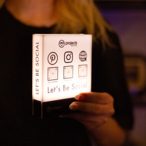May include: A rectangular, illuminated sign with the text "Let's Be Social" and social media icons. The sign features logos for Pinterest, Instagram, and a globe. The sign is held by a person.