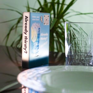 May include: A rectangular illuminated sign with the text "Already thirsty?" and an image of a blue cocktail with a lemon slice. The sign also includes a QR code and the phrase "Order your drink here!"