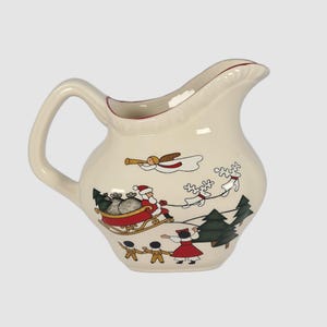 May include: A white ceramic creamer with a Christmas scene featuring Santa Claus in a sleigh pulled by reindeer, angels, and children. The creamer has a handle and a spout.