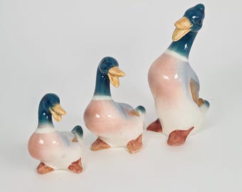 May include: Three ceramic duck figurines in varying sizes. The ducks have blue heads, yellow beaks, and pink and white bodies. The ducks are posed in different positions, with one duck looking up.
