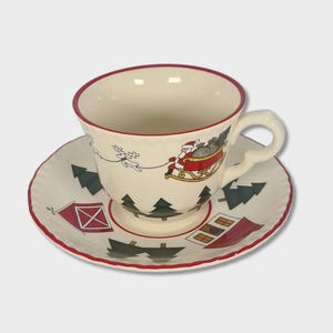 May include: A white ceramic teacup and saucer set with a Christmas theme. The cup and saucer feature a red trim and a festive design of Santa Claus in a sleigh pulled by reindeer, along with houses and pine trees.