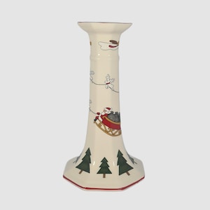May include: White ceramic candle holder with a Christmas scene of Santa Claus in a sleigh pulled by reindeer. The base of the holder is octagonal and features green pine trees.
