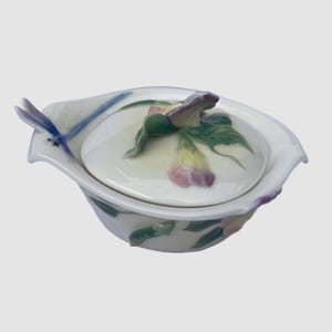 May include: A white ceramic trinket dish with a lid, featuring a floral design in shades of purple, green, and yellow. A blue dragonfly decorates the side. The dish is ideal for storing small jewelry items.