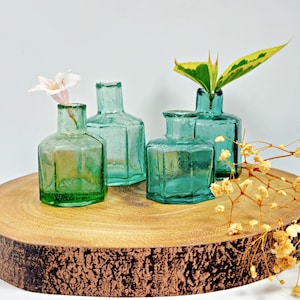 May include: Four teal blue glass bottles with a textured surface, arranged on a wooden slice. The bottles are different sizes and shapes. A small pink flower is in one of the bottles. A sprig of green leaves is in another bottle.