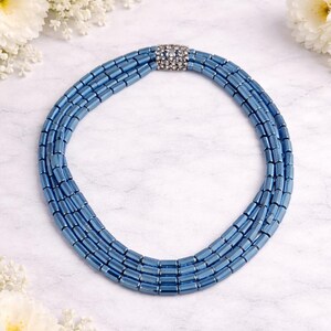 May include: A blue beaded necklace with a silver clasp. The necklace is composed of multiple strands of small, rectangular beads. The clasp is adorned with small, clear stones. The necklace is displayed on a white surface with white flowers.