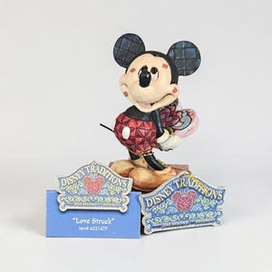 May include: A Mickey Mouse figurine from the Disney Traditions collection. The figurine features Mickey in a red and black outfit, holding a heart-shaped item. The base includes two signs that read "Disney Traditions" and "Love Struck".