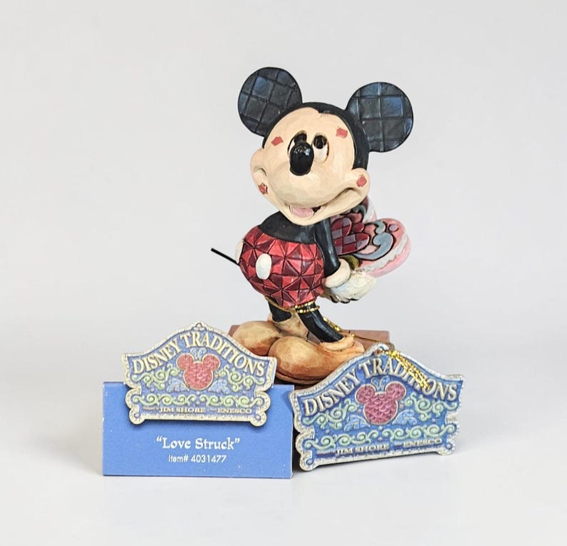 May include: A Mickey Mouse figurine from the Disney Traditions collection. The figurine features Mickey in a red and black outfit, holding a heart-shaped item. The base includes two signs that read "Disney Traditions" and "Love Struck".