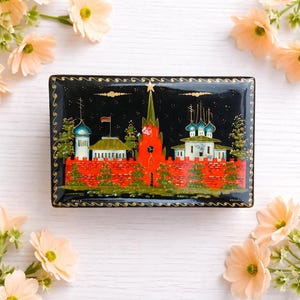 May include: A rectangular black lacquer box with a detailed painting of a cityscape featuring red brick walls, red towers, and green-roofed buildings. The box is decorated with gold trim and a night sky background.