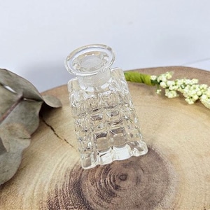 May include: A clear glass bottle with a square, textured design. The bottle has a wide mouth and a clear glass stopper.