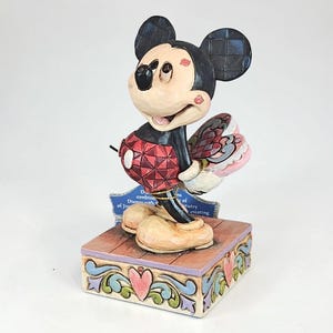 May include: A Mickey Mouse figurine with a red and black outfit, holding a pink and red fan. The figurine stands on a decorative wooden base with a blue plaque. The base has a heart and scroll design.