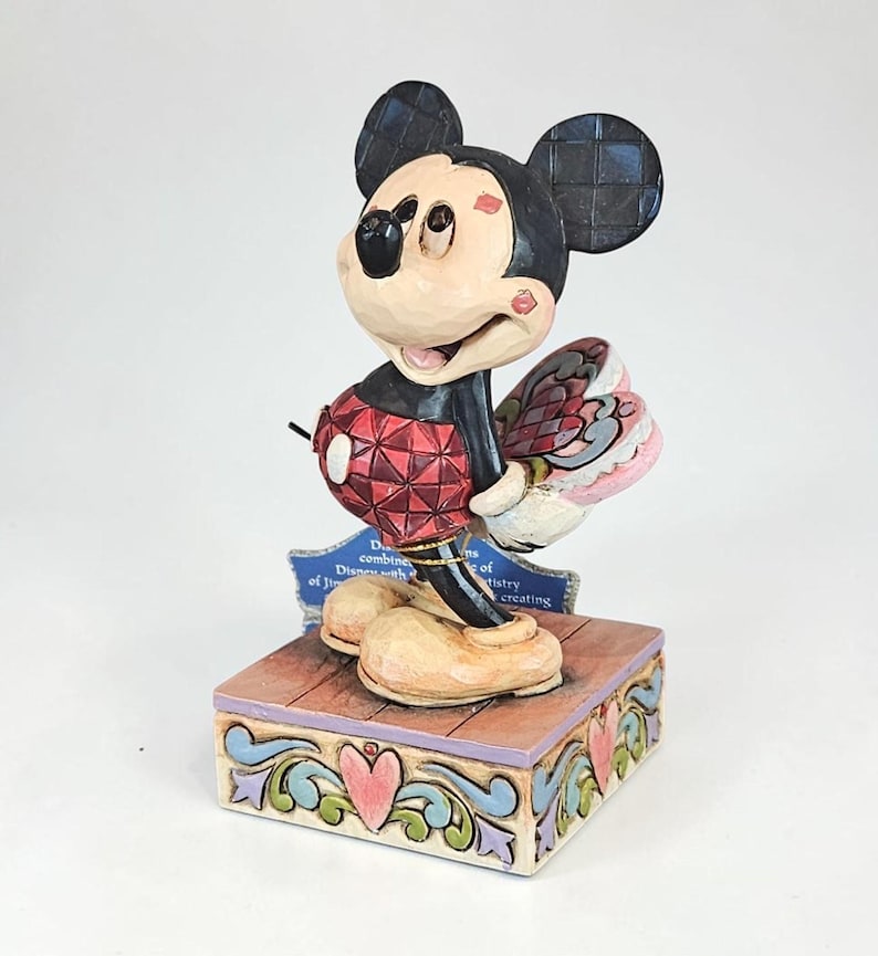 May include: A Mickey Mouse figurine with a red and black outfit, holding a pink and red fan. The figurine stands on a decorative wooden base with a blue plaque. The base has a heart and scroll design.