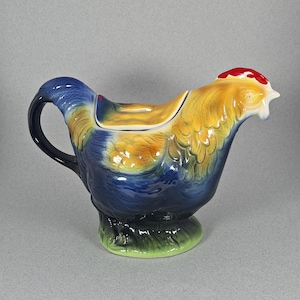 May include: A ceramic rooster-shaped jug with a handle, spout, and lid. The jug features a vibrant colour scheme, with blue, yellow, and red hues. The base is green, and the jug is designed to resemble a rooster.