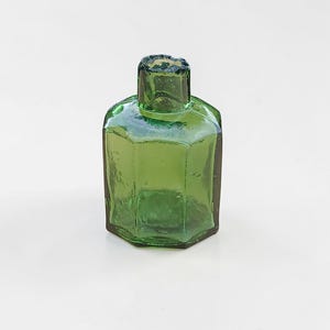 Peut inclure: Petite bouteille en verre vert antique avec un corps hexagonal et un col carré. La bouteille a une surface légèrement texturée et est vide. La bouteille est probablement utilisée pour les parfums ou les médicaments.