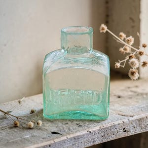 May include: A vintage, rectangular, pale-green glass bottle with the embossed word "JEFFELD". The bottle has a square neck and is displayed on a weathered wooden surface. Dried flowers are in the background.