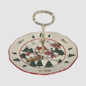 May include: A white ceramic serving tray with a red rim and a gold handle. The tray features a Christmas scene with a house, trees, and people riding in a sleigh.