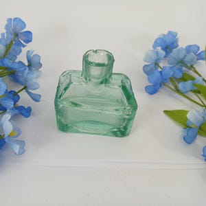 May include: A small, clear green glass ink bottle with a square base and a round opening. The bottle is sitting on a white surface.