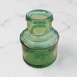 May include: A small, antique green glass bottle with a rounded base and a short, cylindrical neck. The bottle has a slightly iridescent quality and is likely made of pressed glass. The bottle is approximately 3 inches tall.