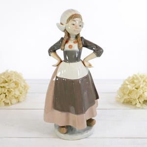 May include: A ceramic figurine of a young girl in traditional Dutch attire. The figurine features a white bonnet, a gray and white dress with a brown skirt, and brown shoes. The girl has her hands on her hips and is standing on a round base.