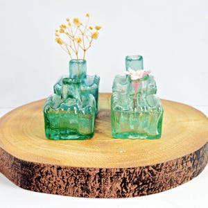 Peut inclure: Deux petits vases carrés en verre bleu-vert avec des surfaces texturées, posés sur une tranche de bois. Un vase contient des fleurs séchées et l'autre une seule fleur rose.