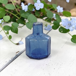 May include: A small, antique cobalt blue glass bottle with a hexagonal body and a short, rectangular neck. The bottle is displayed on a white wooden surface, with green leaves and small blue flowers in the background. The bottle is approximately 3 inches tall.