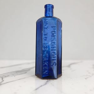May include: A deep blue antique poison bottle with the embossed text "POISONOUS NOT TO BE TAKEN". The bottle has a rectangular shape with vertical ribbed sides and a rounded neck.