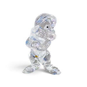 May include: A clear, faceted crystal figurine depicting a character with a large head, prominent nose, and crossed arms. The figure has a smooth, reflective surface that catches light, creating prismatic highlights across its sculpted form.