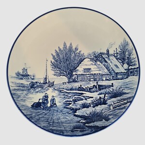 May include: A round, white ceramic plate with a blue border. The plate features a detailed blue and white illustration of a rural winter scene with a windmill, boat, and thatched-roof houses.