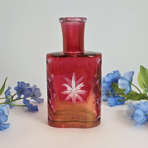May include: A rectangular ruby red glass bottle with a cylindrical neck. The bottle has a starburst design etched into the glass. The bottle is surrounded by blue flowers.