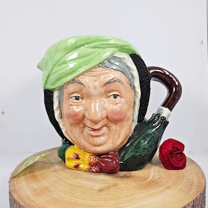 May include: A ceramic character mug with a green hat, a brown handle, and a smiling face. The mug is decorated with a green and brown pattern.