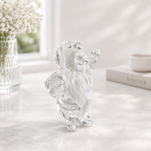 Peut inclure: Figurine transparente en cristal représentant un ours avec une longue barbe et les bras tendus. L'ours est facetté, captant la lumière et créant un effet scintillant. La figurine est exposée sur une surface blanche, avec un vase de fleurs blanches en arrière-plan.
