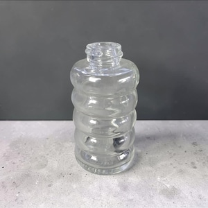 May include: A clear glass bottle with a unique, stacked, bubble-like design. The bottle has a rounded base and a narrow neck with a threaded opening. The transparent glass allows a clear view of the contents. The bottle is suitable for storing liquids.