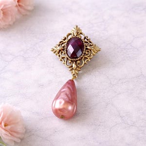 May include: A vintage-style brooch with a gold-tone filigree setting. The brooch features a faceted, deep red oval gemstone at the center, and a pear-shaped, pink pearl drop. The brooch is an elegant accessory.