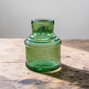 May include: An antique green glass bottle, likely an ink bottle, with a rounded base and cylindrical neck. The bottle is made of transparent green glass and sits on a wooden surface. The bottle has a textured surface.